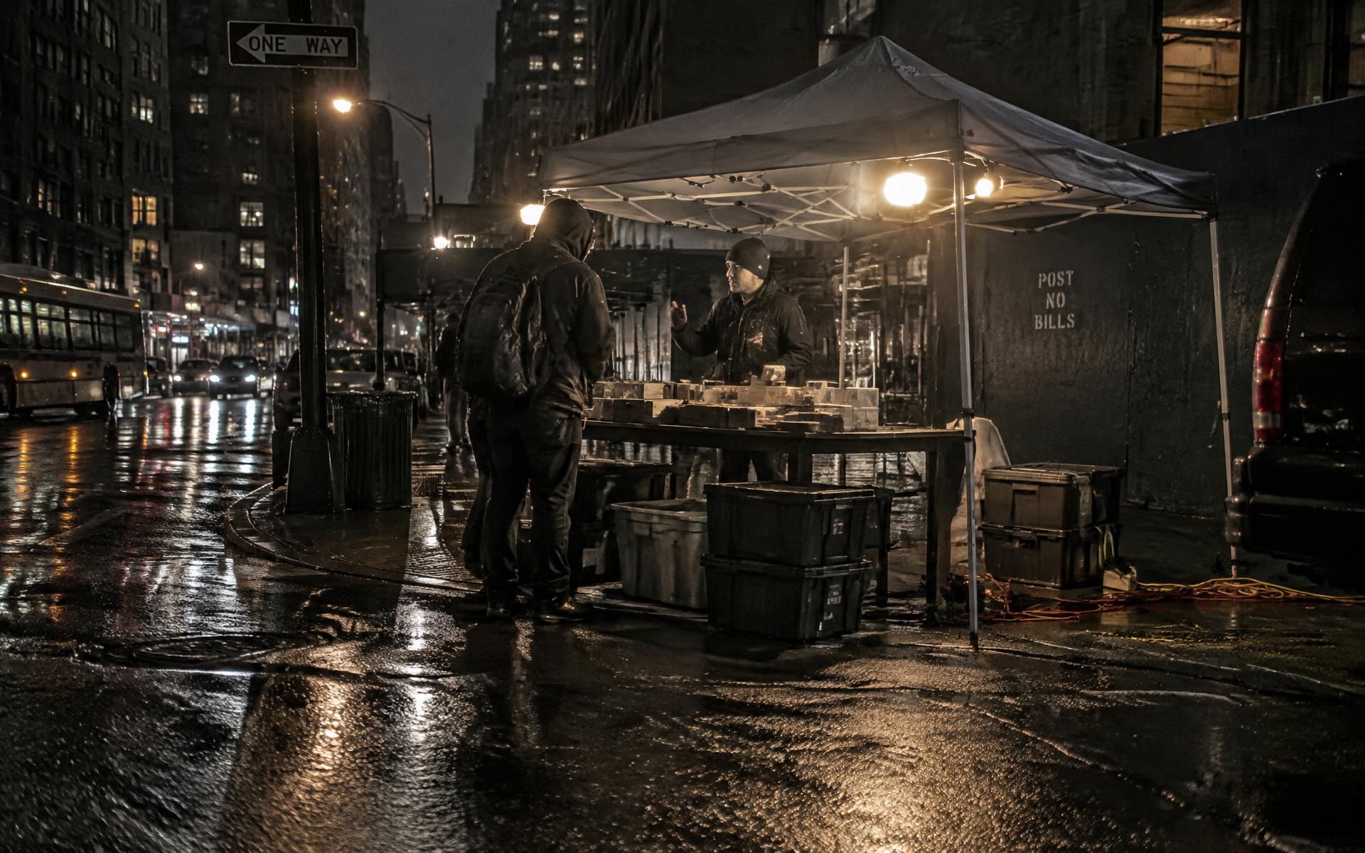 An unbranded pop-up sales stand under work lights on a wet New York street at night, a representative talking with a passer-by